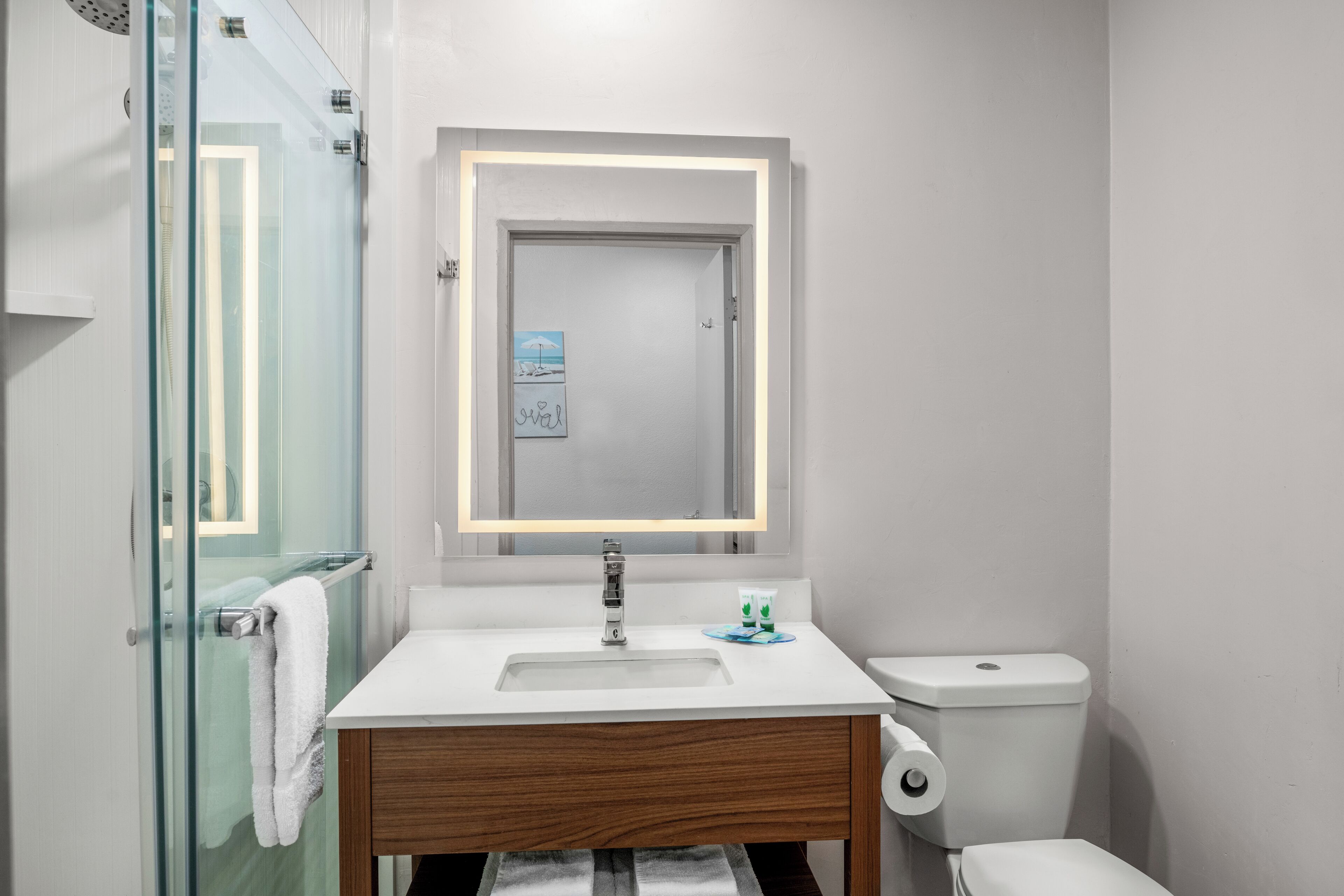 A modern bathroom with a glass shower enclosure, a white sink with a wooden vanity, a toilet, and a framed artwork above the mirror.