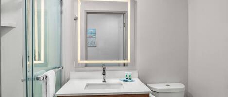 A modern bathroom with a glass shower enclosure, a white sink with a wooden vanity, a toilet, and a framed artwork above the mirror.