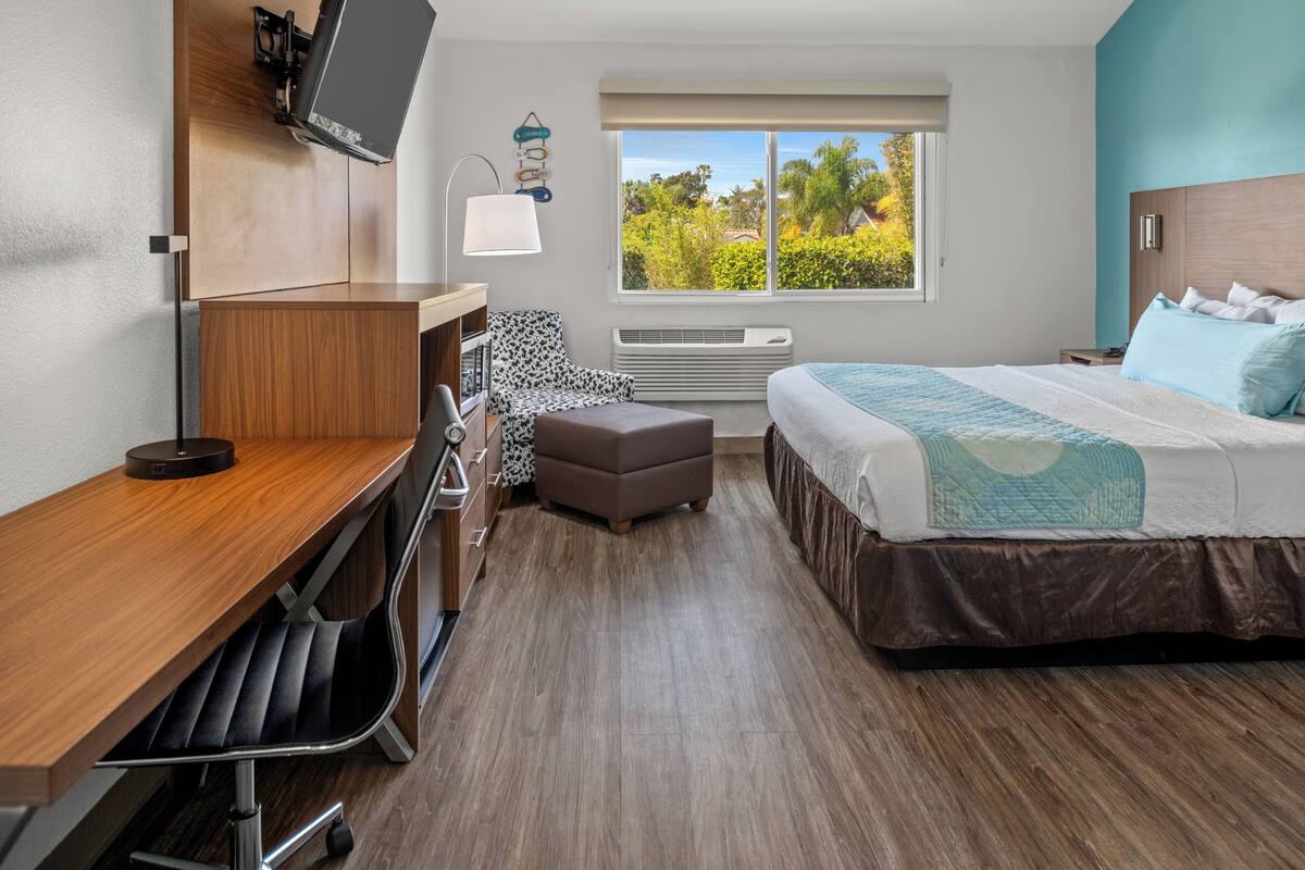 Habitación de hotel con escritorio de madera, cama con colcha azul y blanca, un pequeño reposapiés, una silla, televisor montado en la pared y ventana con vista a follaje.