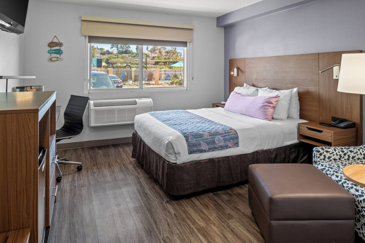 Habitación de hotel con cama, mesita de noche, lámpara y vistas al exterior.