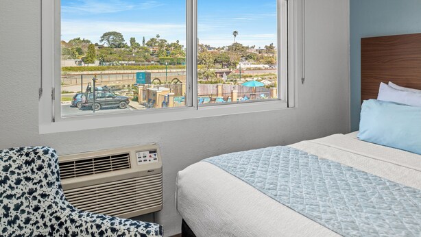 Un dormitorio con cama, una silla y una ventana que da a un estacionamiento y edificios.