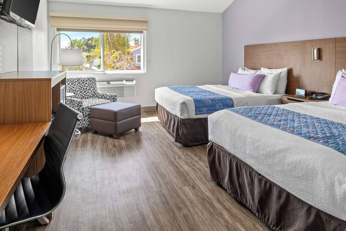 Habitación de hotel con dos camas, un escritorio, una silla y ventana con vistas a árboles y edificios.