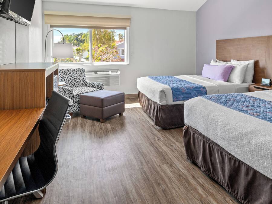 Habitación de hotel con dos camas, un escritorio, una silla y ventana con vistas a árboles y edificios.