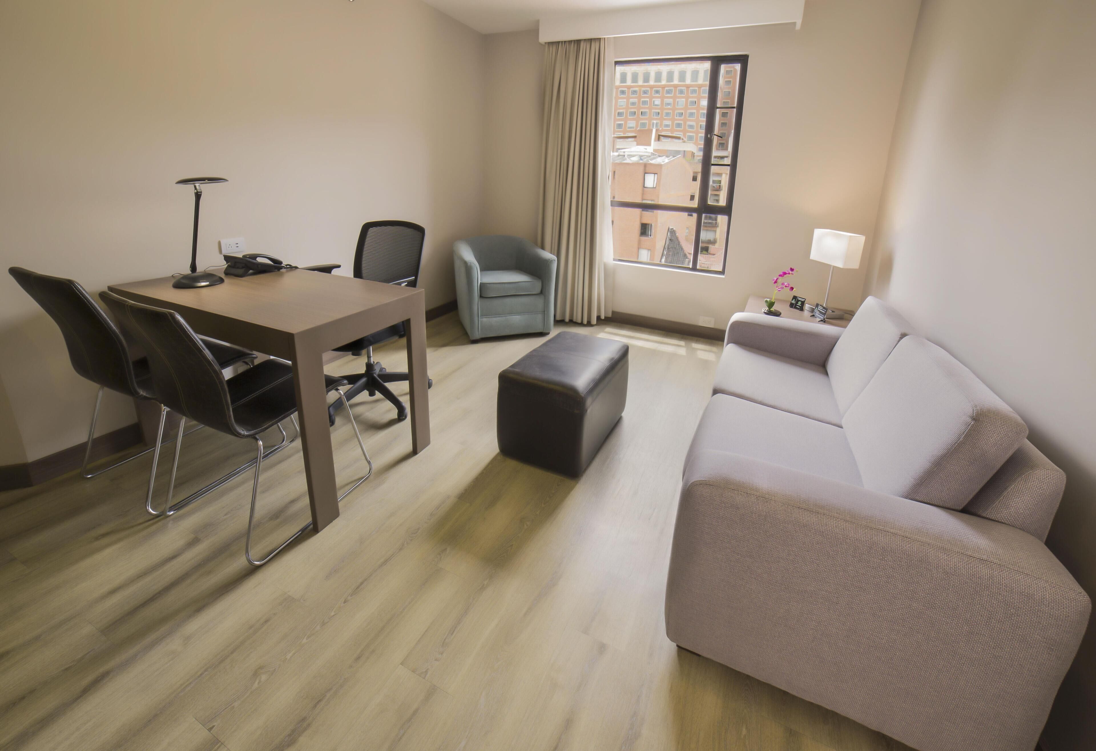 A hotel room with a dining table, two chairs, a sofa, a small ottoman, a lamp, and a window with curtains.