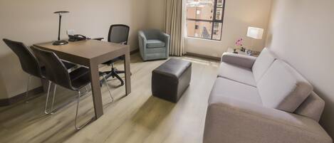 A hotel room with a dining table, two chairs, a sofa, a small ottoman, a lamp, and a window with curtains.