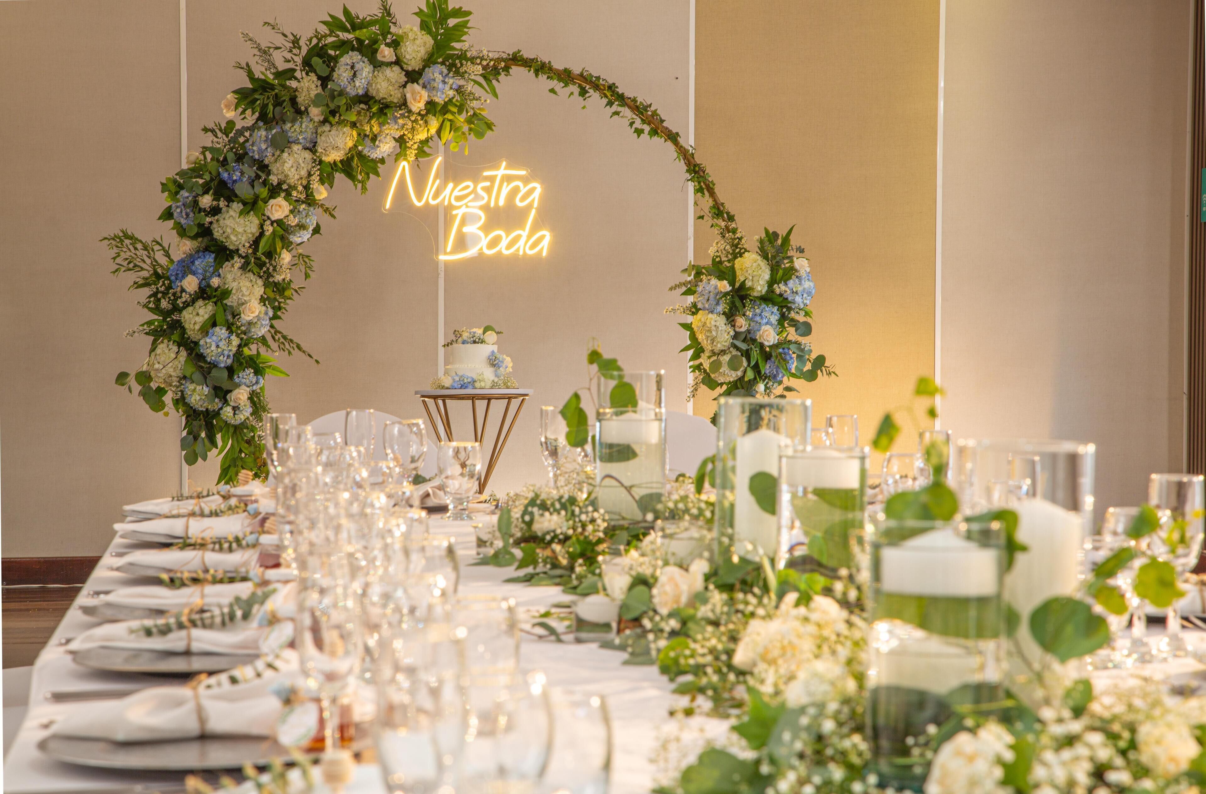 A banquet table set with glassware and floral centerpieces, a neon sign reading 'Nuestra Boda,' and a cake on a stand.