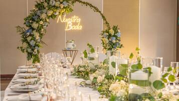 A banquet table set with glassware and floral centerpieces, a neon sign reading 'Nuestra Boda,' and a cake on a stand.