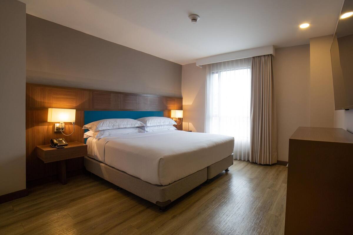 Ein Hotelzimmer mit einem Bett, Nachttischlampen, einem Schreibtisch mit Telefon, einem Flachbildfernseher und einer Tür.