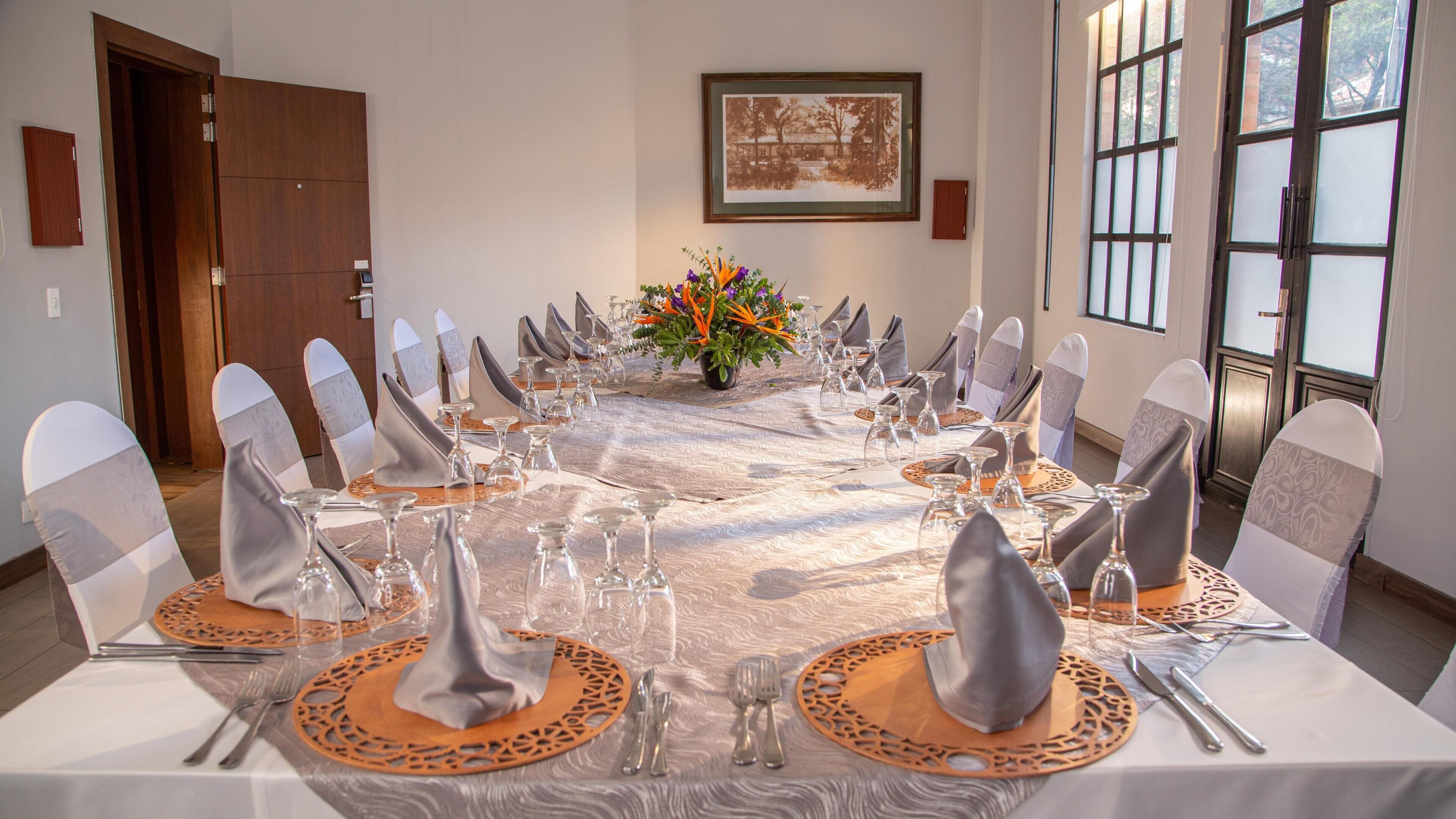 Un salón de conferencias con una larga mesa dispuesta para una reunión, sillas con fundas blancas y grises, y un cuadro en la pared.