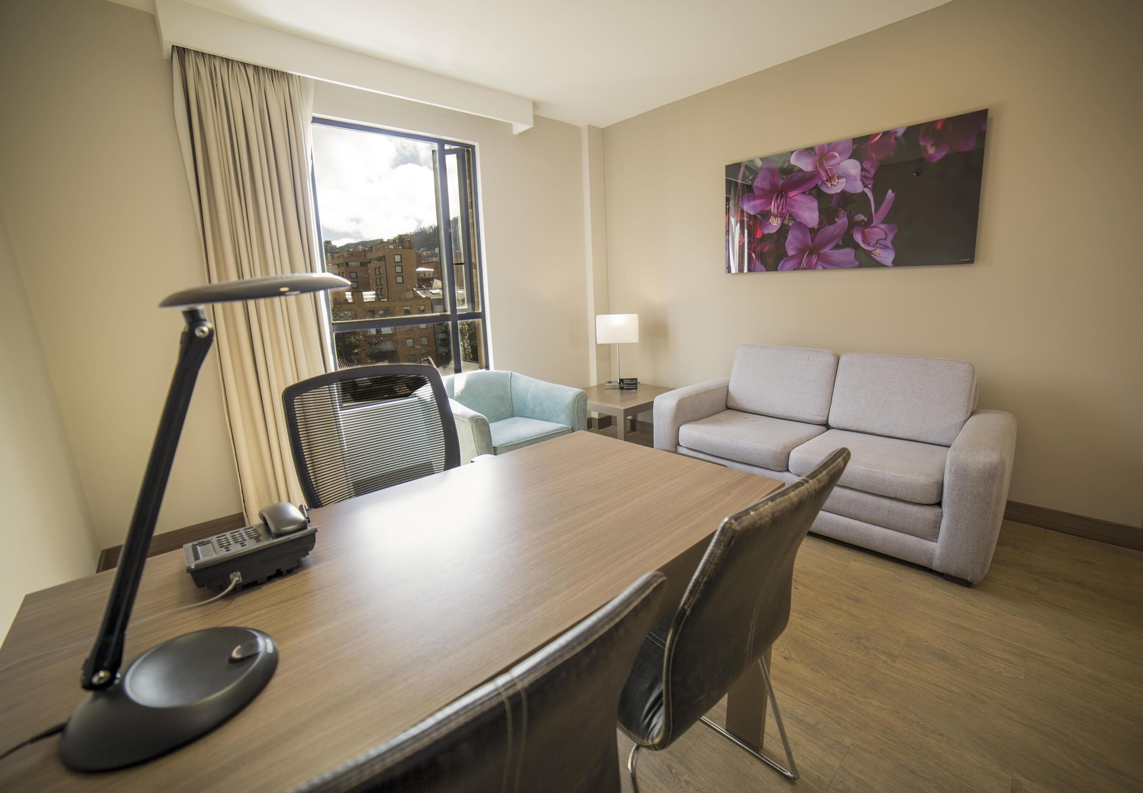 Habitación de hotel con escritorio de madera, lámpara, silla, sofá, un cuadro y ventana con cortinas.