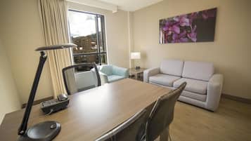 Habitación de hotel con escritorio de madera, lámpara, silla, sofá, un cuadro y ventana con cortinas.