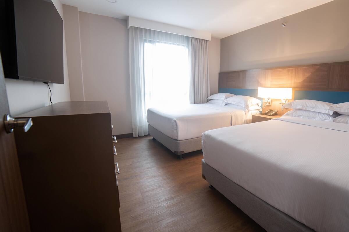 Ein Hotelzimmer mit zwei Betten, einem Schreibtisch, einem Fernseher und einem Fenster mit Vorhängen.