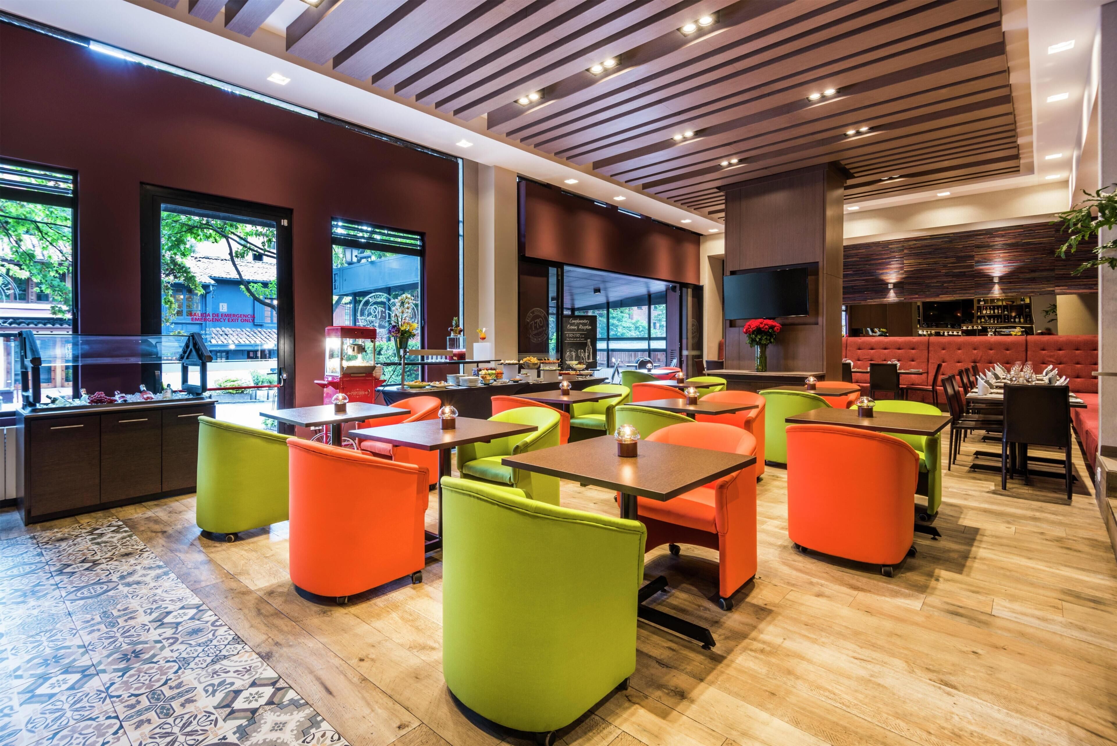 A modern restaurant with a bar area, large windows, and a mix of green and orange seating.