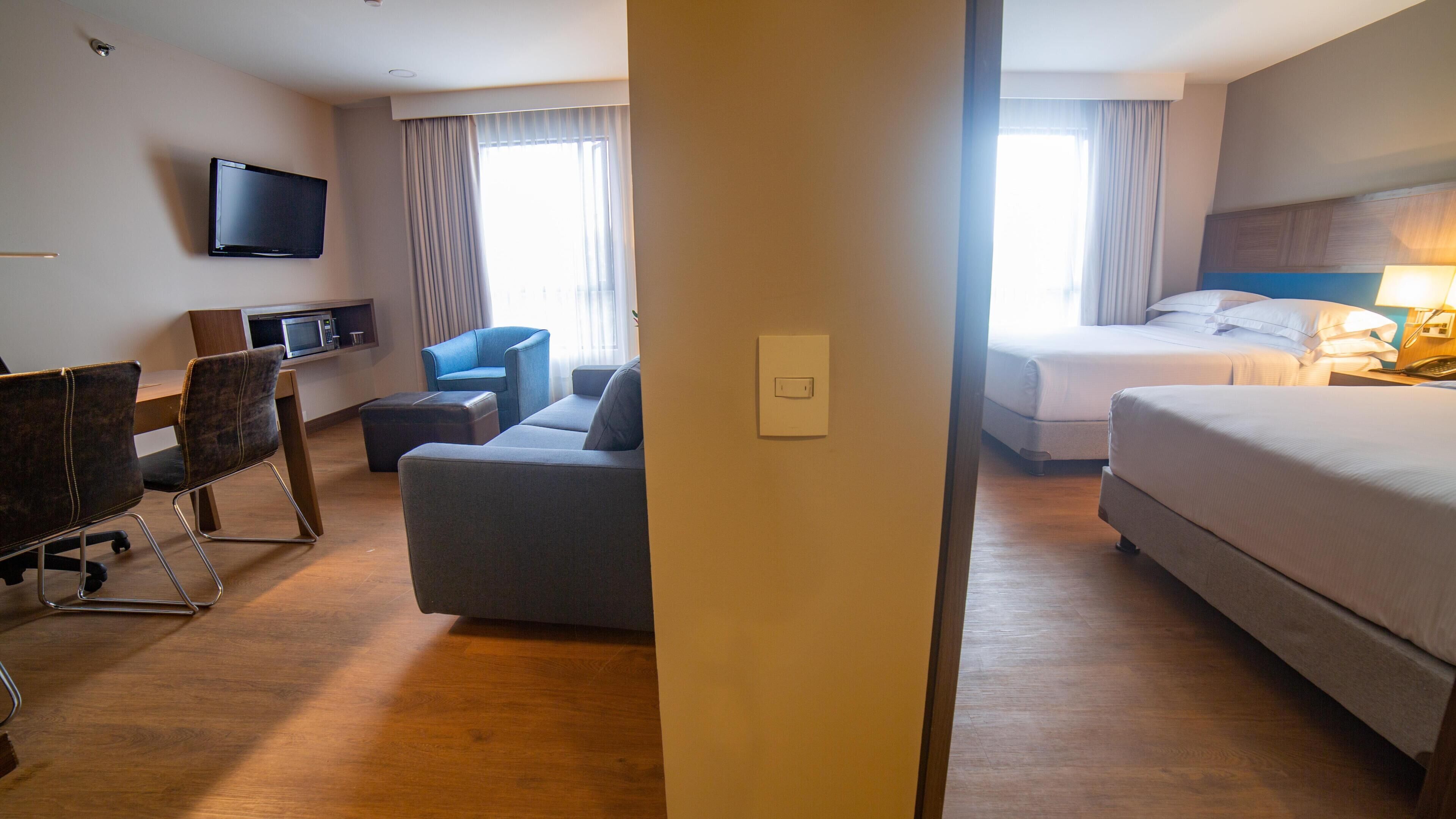 Habitación de hotel con cama, escritorio con silla, sofá, televisor y ventana con cortinas.