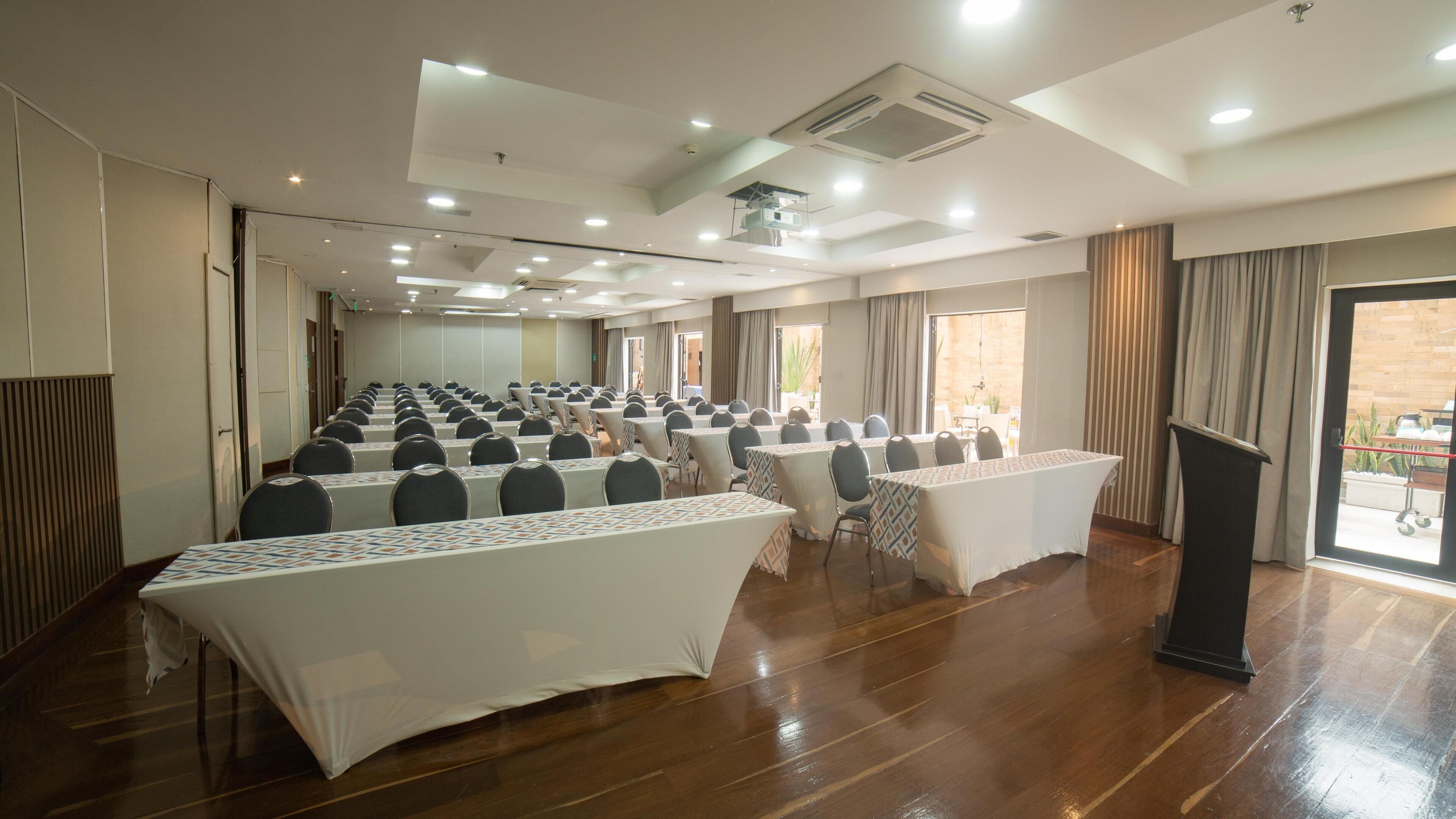 Una sala de conferencias con mesas y sillas dispuestas en filas, ventanas amplias y un atril.