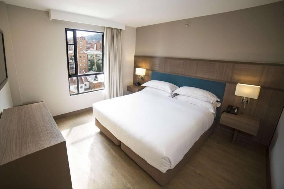 Ein Hotelzimmer mit einem großen Bett, zwei Nachttischen mit Lampen, einem Fenster mit Blick auf Gebäude und einem an der Wand befestigten Flachbildfernseher.