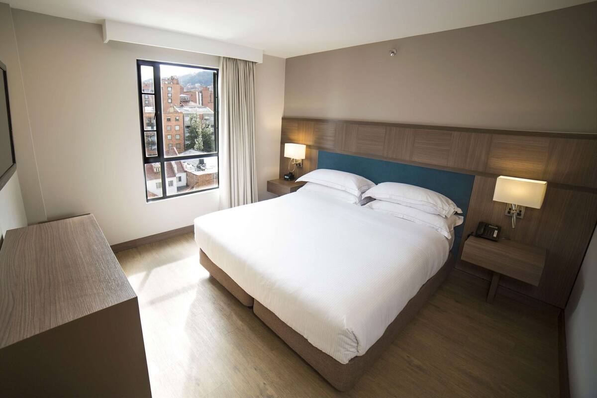 Ein Hotelzimmer mit einem großen Bett, einem Schreibtisch, einem Fenster mit Blick auf die Stadt und einem an der Wand befestigten Fernseher.