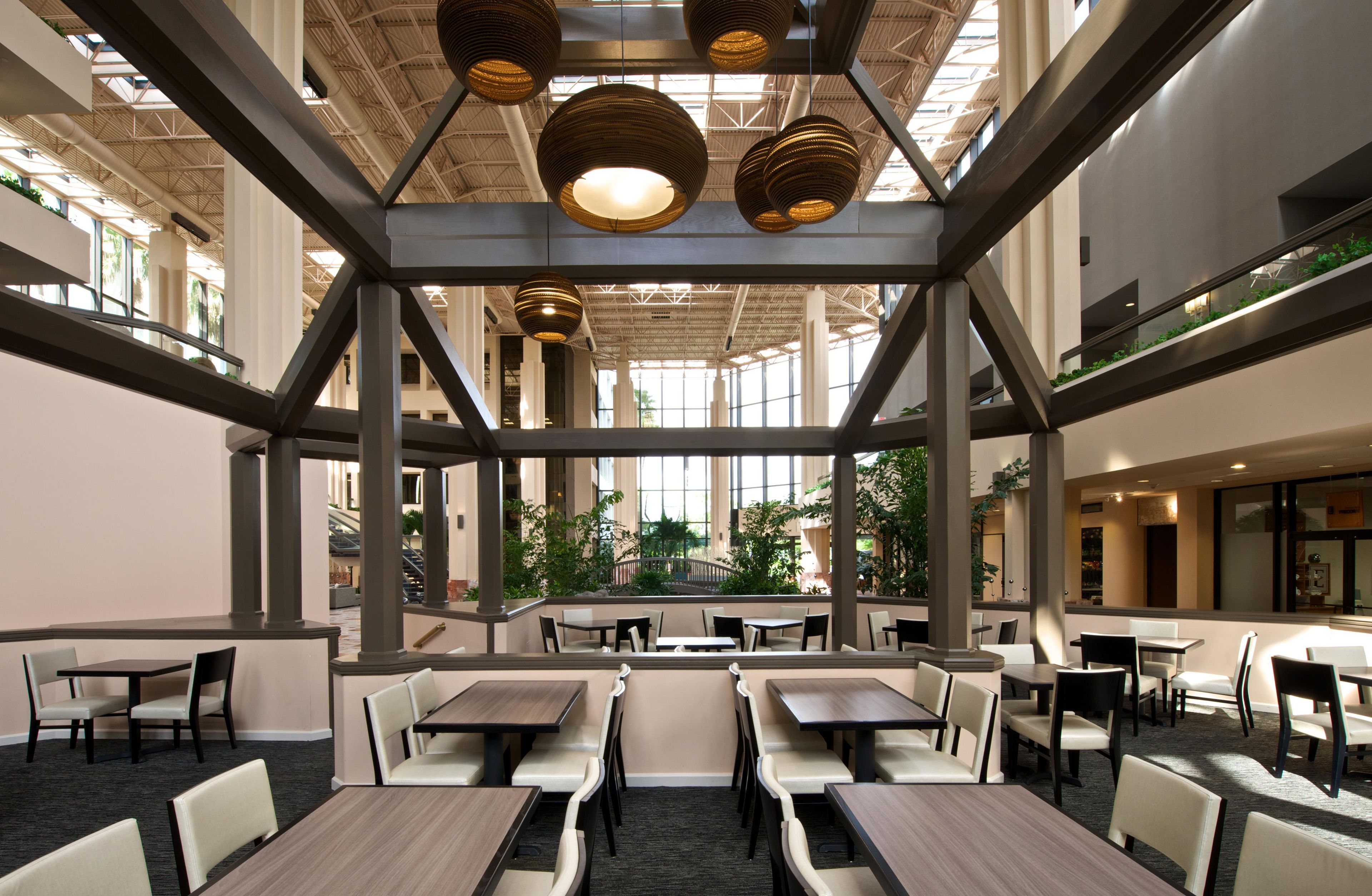 A spacious indoor area with multiple tables and chairs, a high ceiling with hanging lights, and a view of greenery outside.
