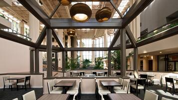 A spacious indoor area with multiple tables and chairs, a high ceiling with hanging lights, and a view of greenery outside.