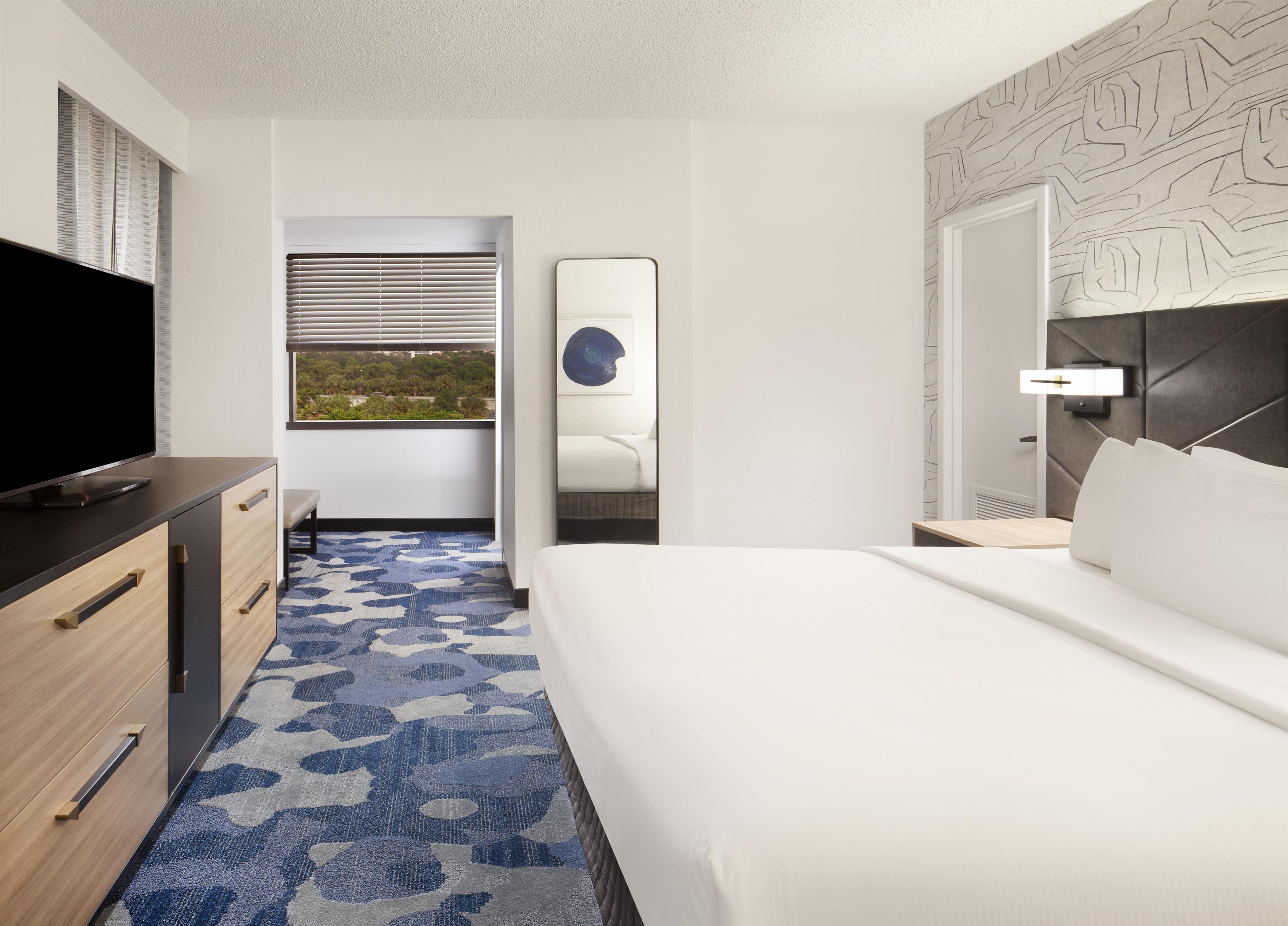 A hotel room with a bed, a TV, a desk, and a window with blinds.