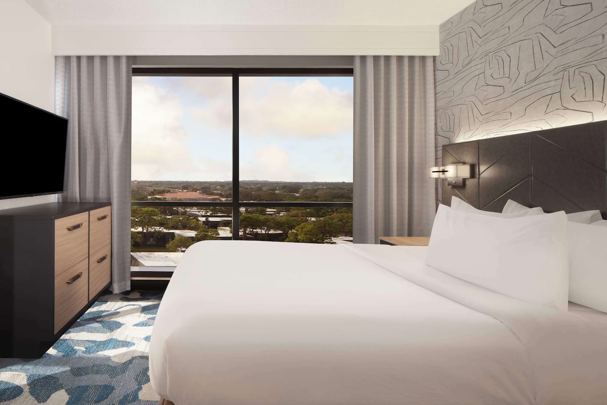 A hotel room with a large bed, a TV, a dresser, and a view of a cityscape through large windows.
