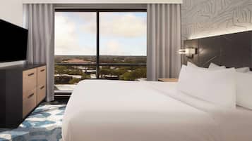 A hotel room with a large bed, a TV, a dresser, and a view of a cityscape through large windows.