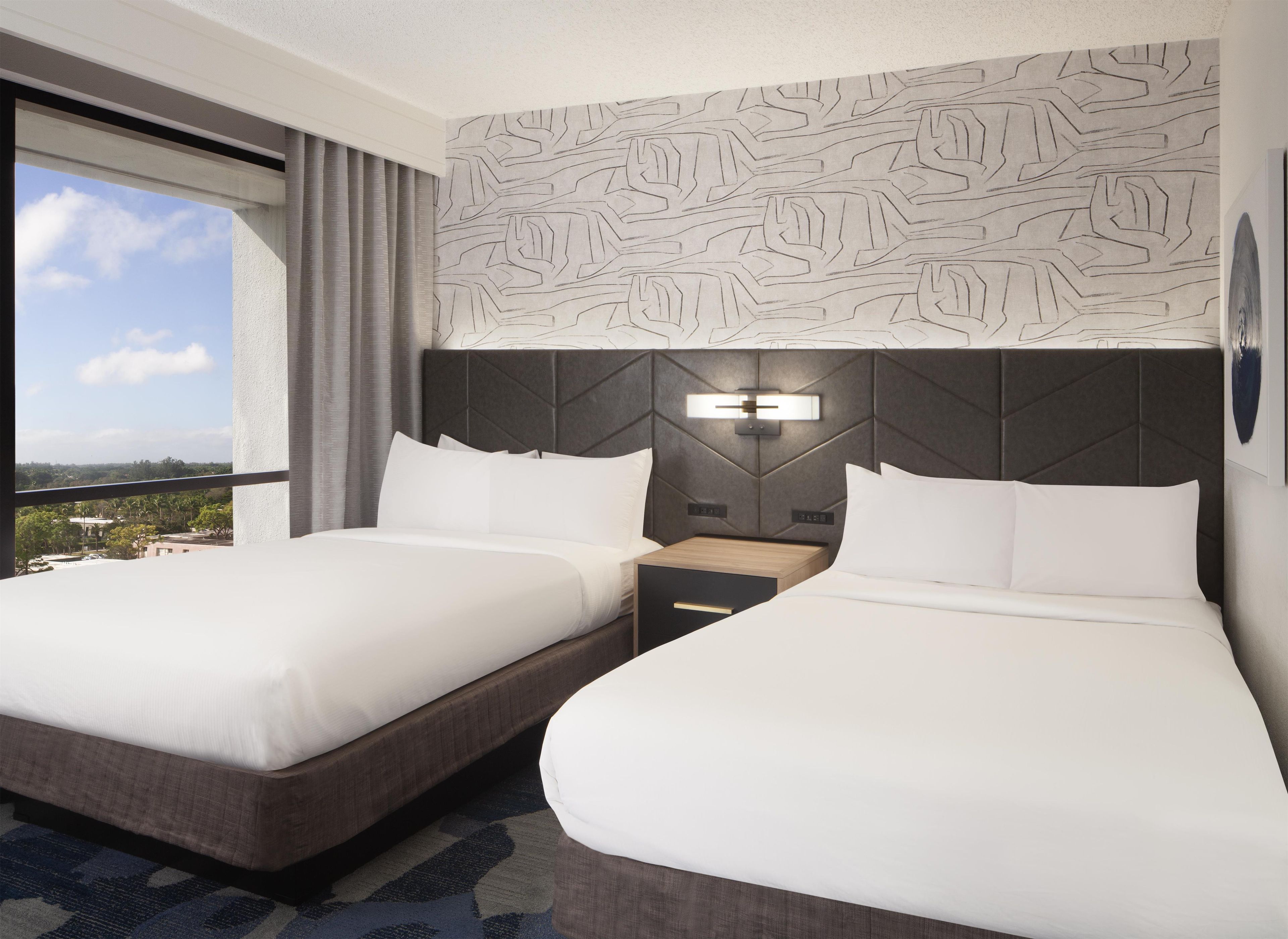 A hotel room with two beds, a patterned wall, a nightstand, and a window with a view.