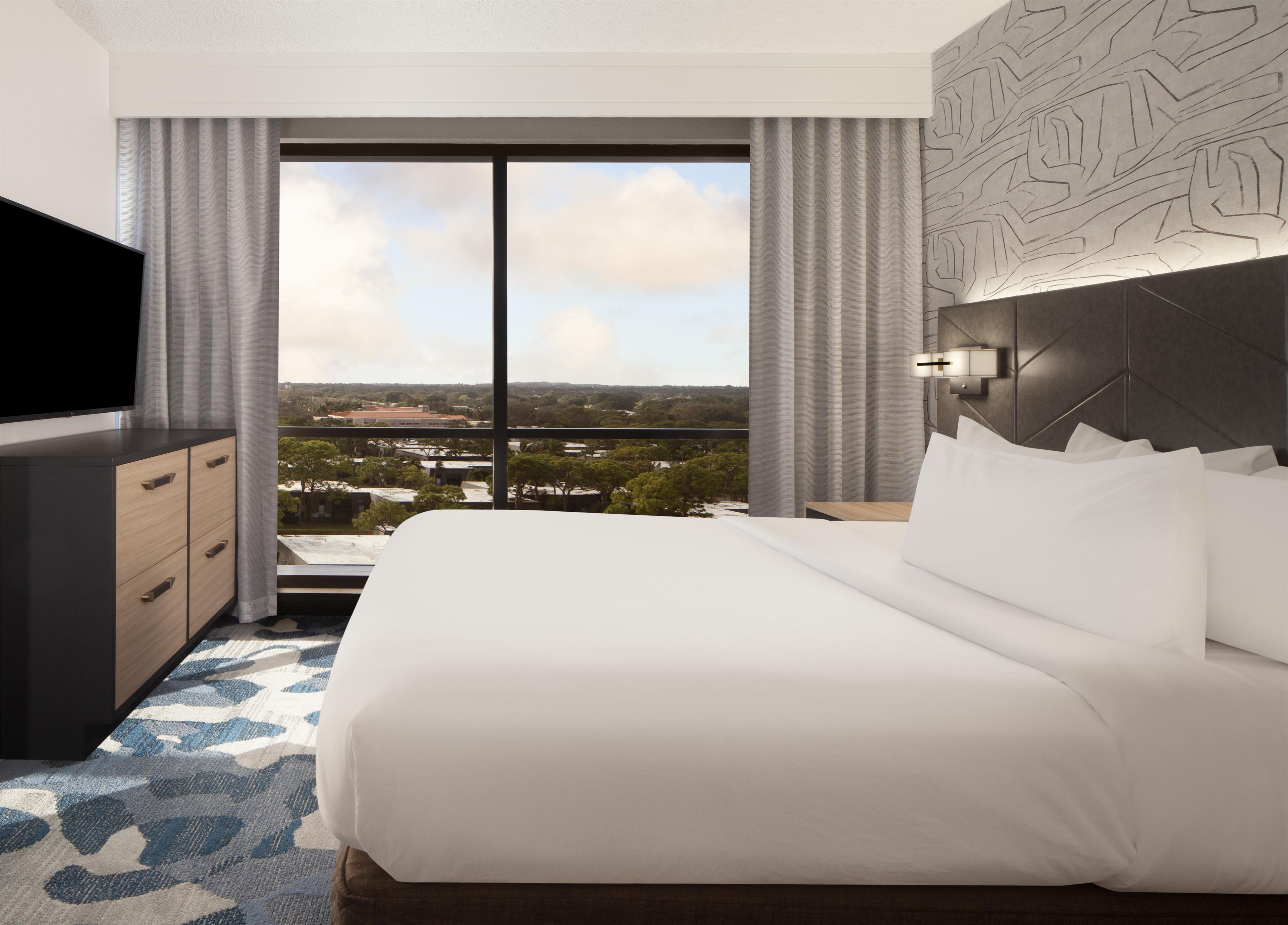 A hotel room with a large bed, a flat-screen TV, a dresser, and a view of a cityscape through a large window.