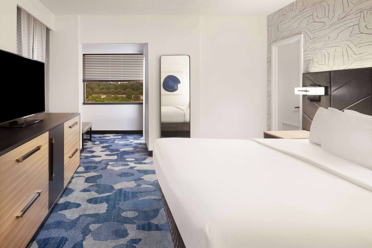 A hotel room with a bed, a TV, a desk, and a window with blinds.