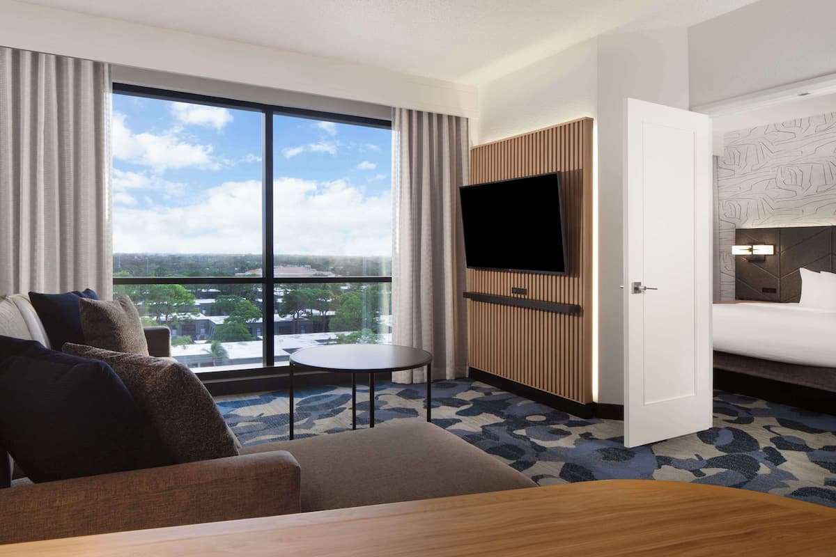 A hotel room with a large window, a flat-screen TV, a bed, a sofa, and a small table.