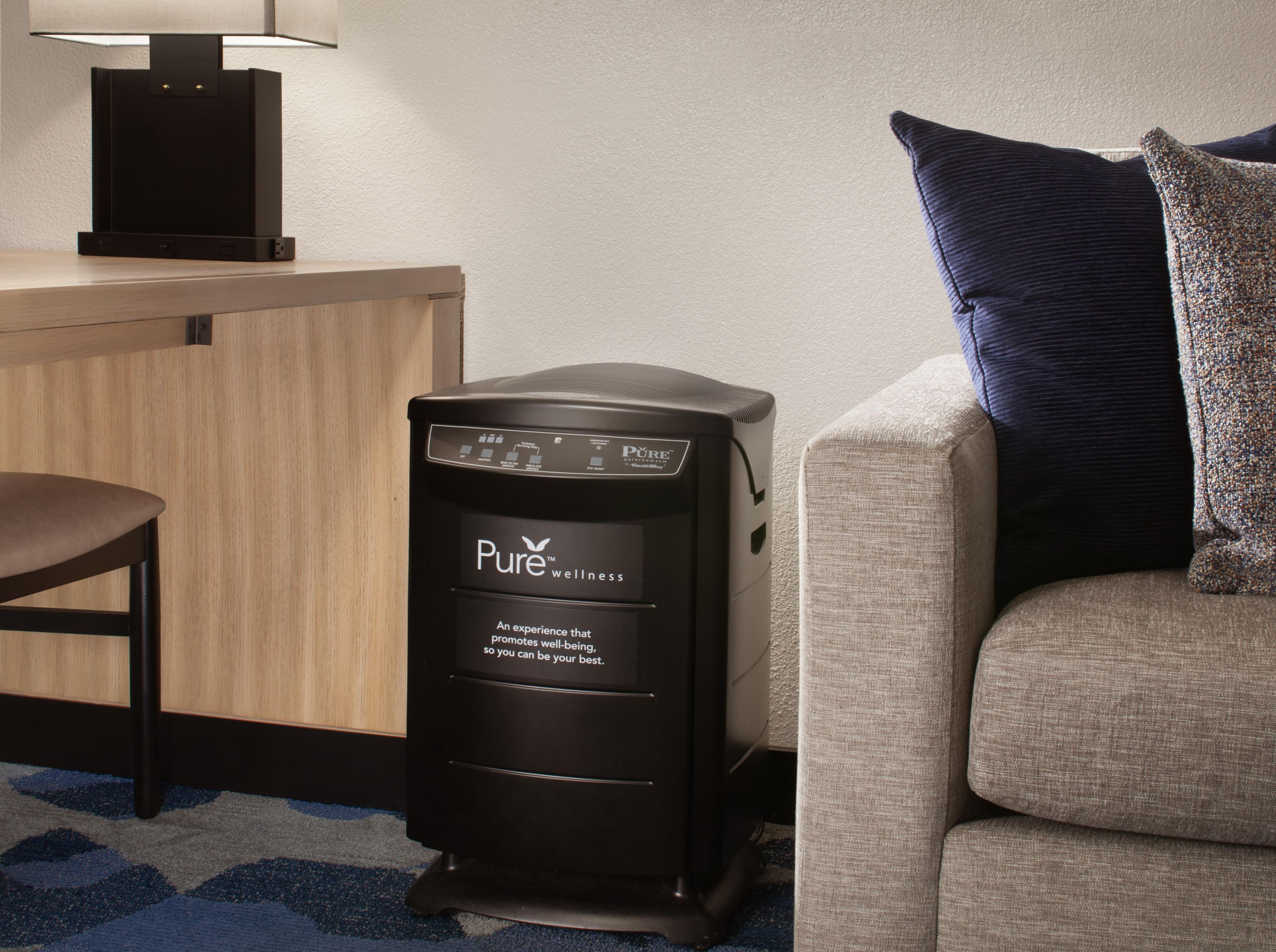 A black wellness station with a control panel and a slogan, a wooden desk with a lamp, and a sofa with a dark pillow.