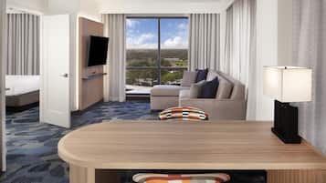 A hotel room with a bed, a sofa, a dining table, a TV, and a view of the city.