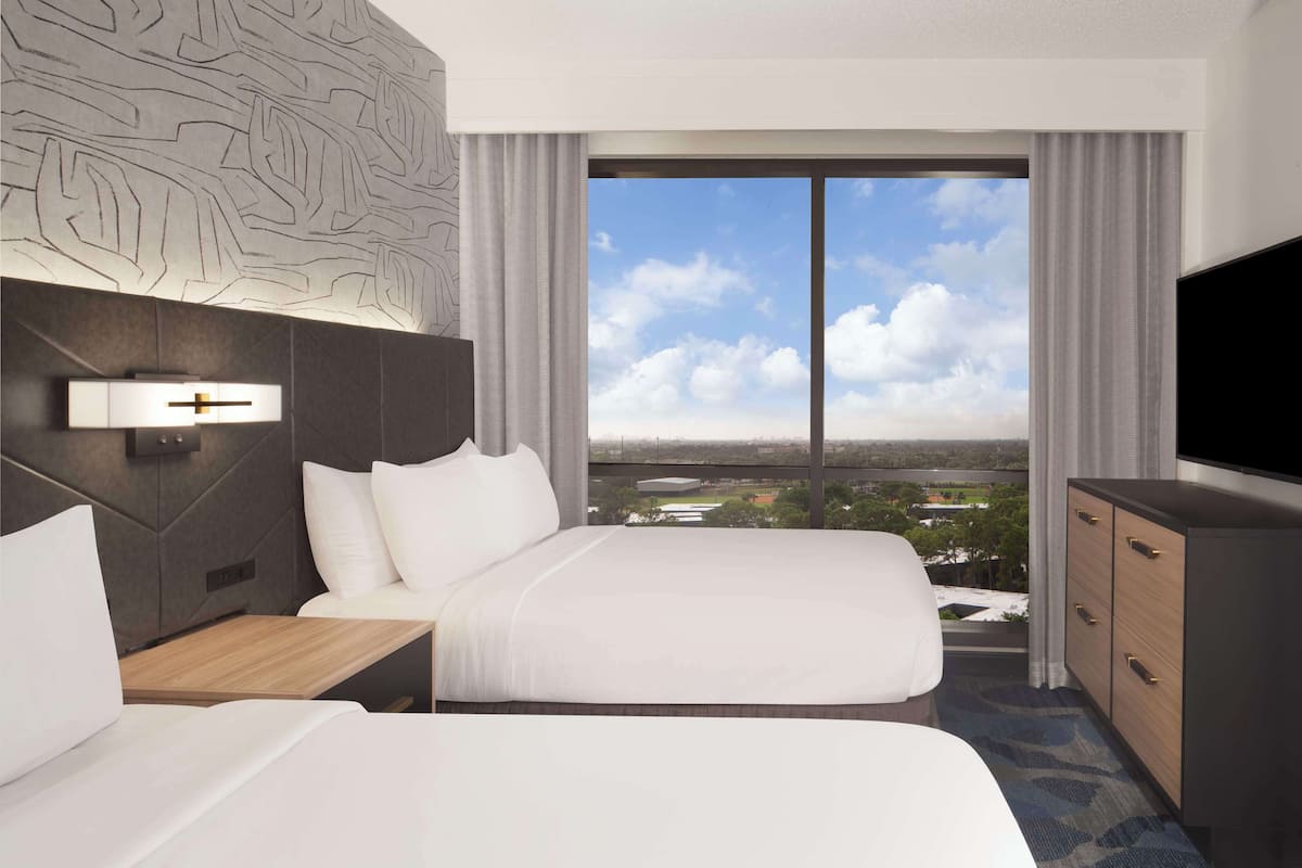 A hotel room with two beds, a TV, a desk, and a view of a cityscape.