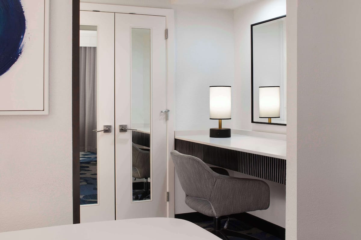 A hotel room with a bed, a desk with a lamp, a chair, a large mirror, and a wardrobe with a sliding door.