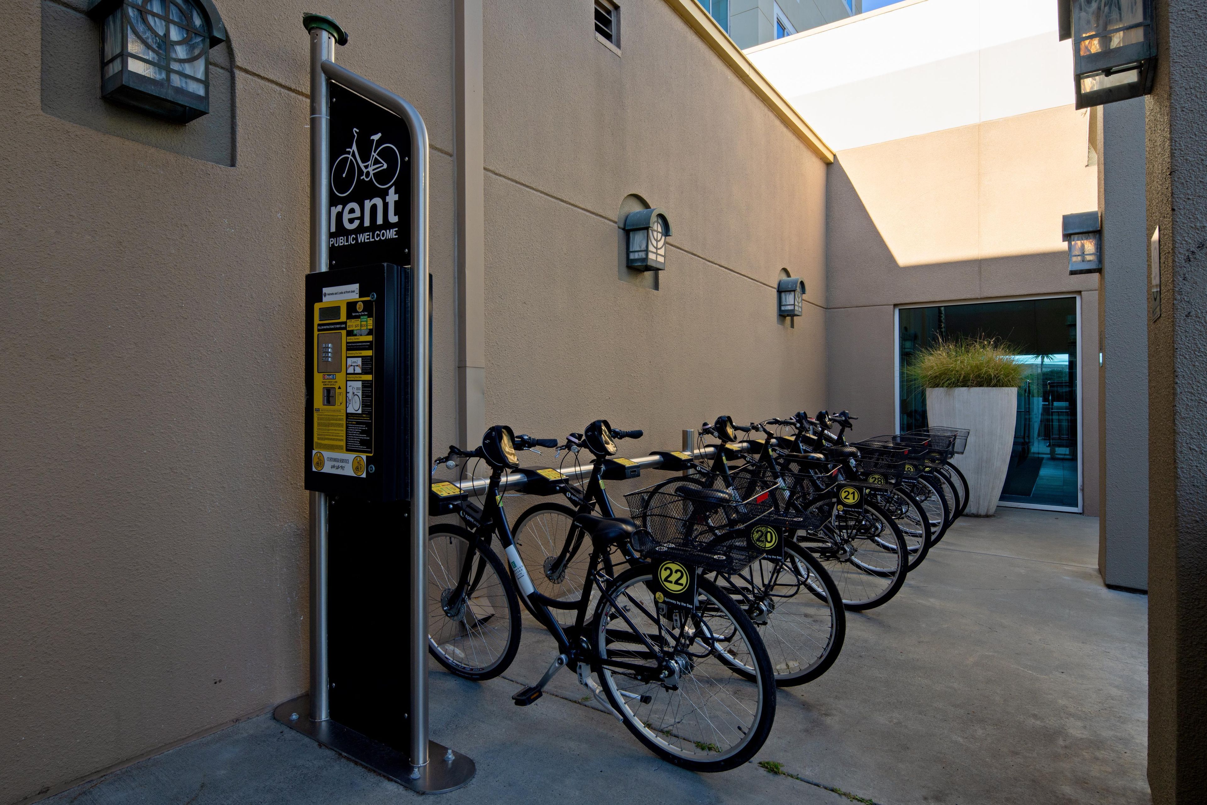 Una estación de alquiler de bicicletas con un cartel que indica que están disponibles, una fila de bicicletas y la entrada de un edificio.