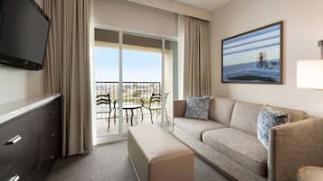 Habitación de hotel con sofá, televisor, banco, mesa de comedor, sillas y un ventanal con vista al océano.