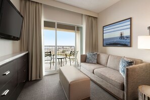 A hotel room with a sofa, a TV, a bench, a dining table, chairs, and a large window with a view of the ocean.