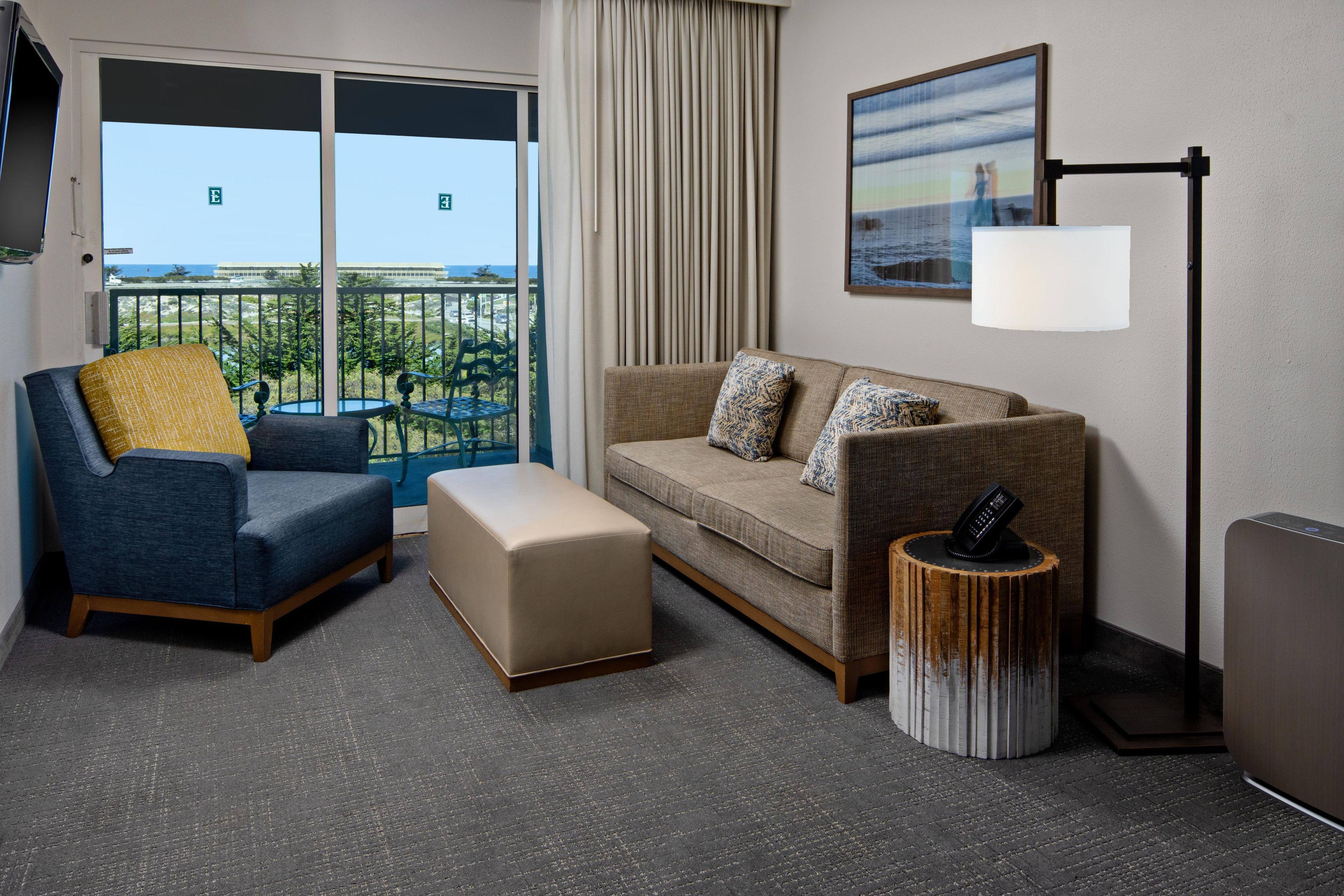 A hotel room with a balcony, a sofa, a chair, a small ottoman, a side table, and a floor lamp.