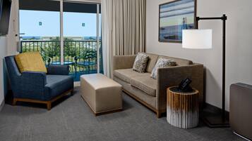 A hotel room with a balcony, a sofa, a chair, a small ottoman, a side table, and a floor lamp.