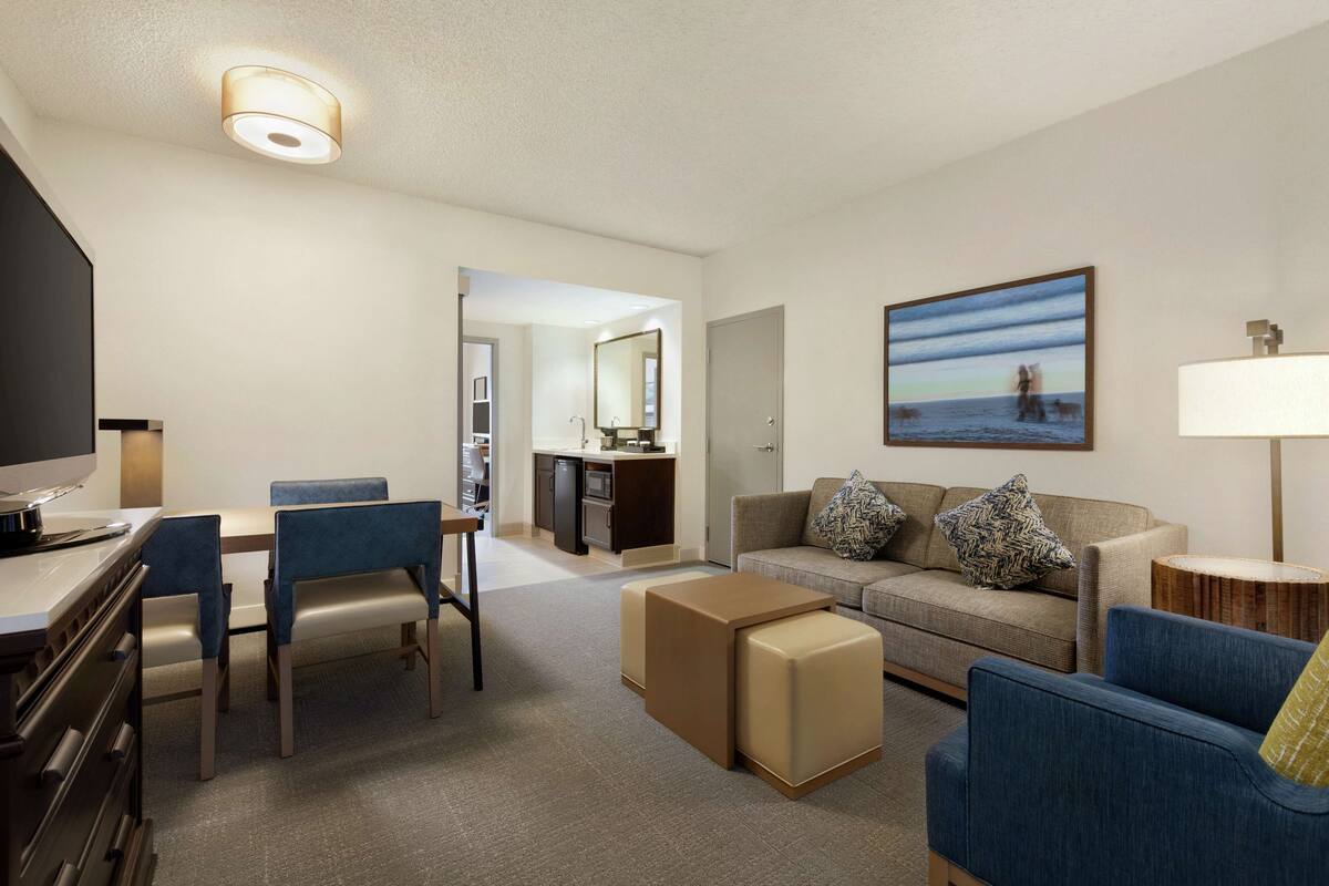 A hotel room with a dining area, a sofa, a TV, and a kitchen in the background.