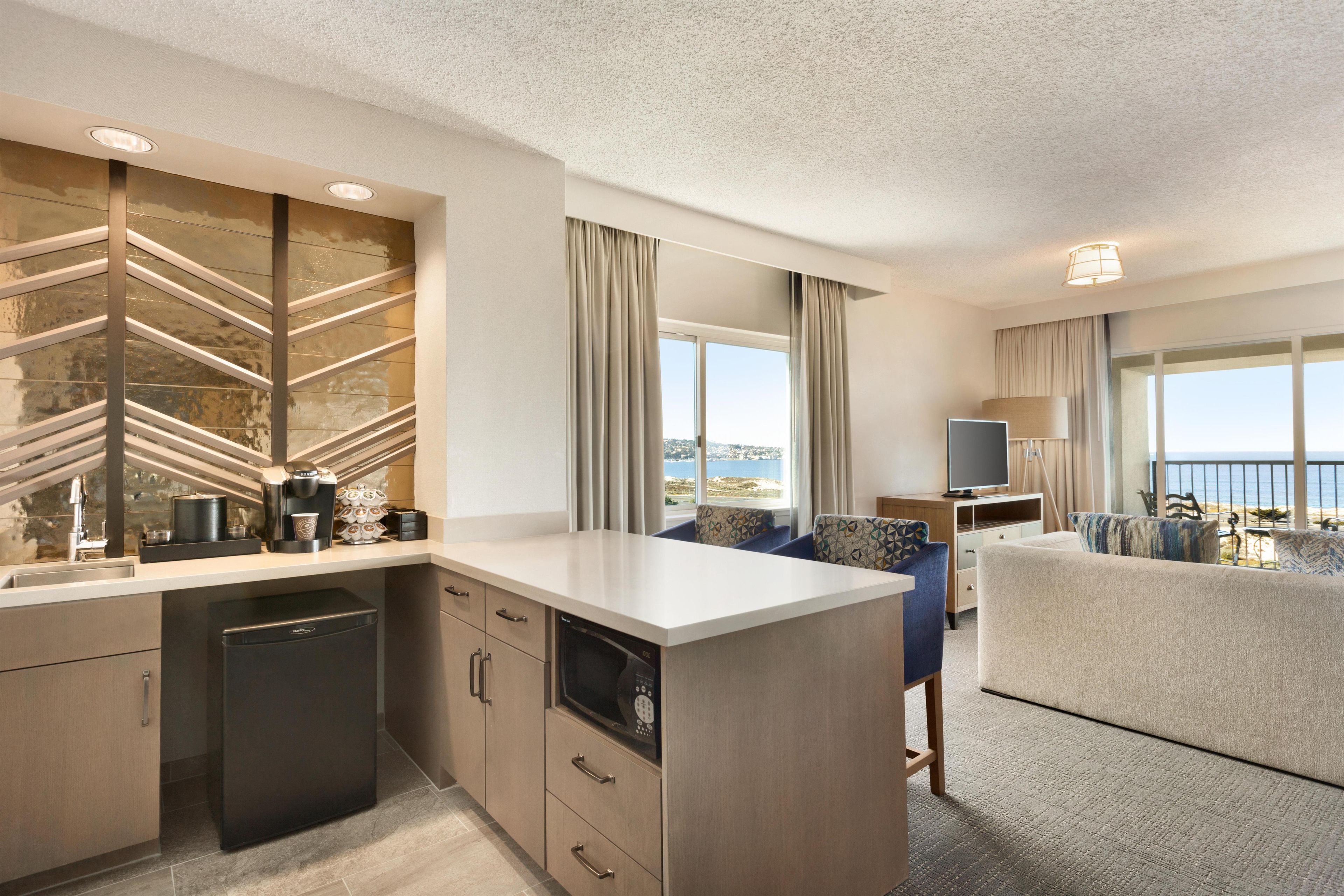 A modern hotel room with a kitchenette, a dining area, and a seating area with a view of the sea.