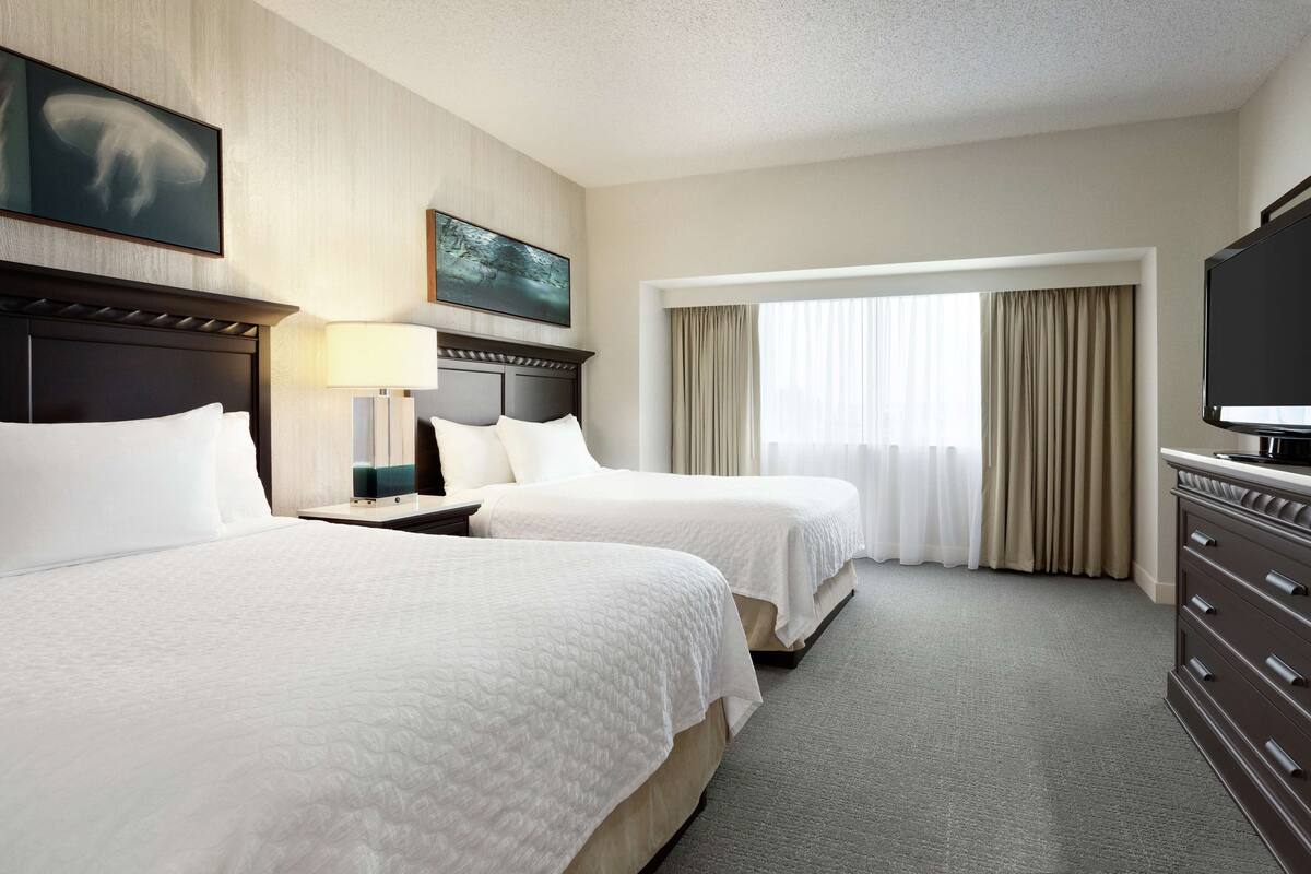 A hotel room with two beds, a TV, a desk, and a window with curtains.