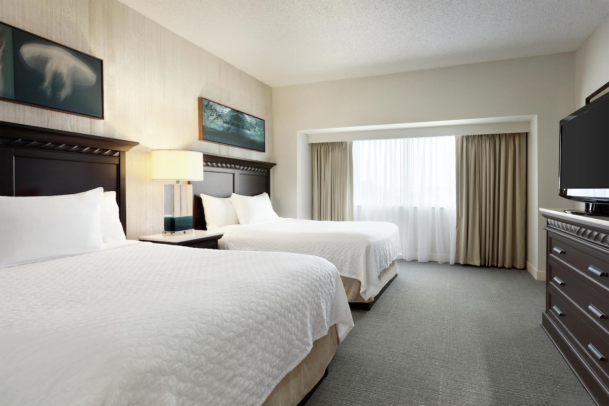 A hotel room with two beds, a TV, a painting, and a window with curtains.