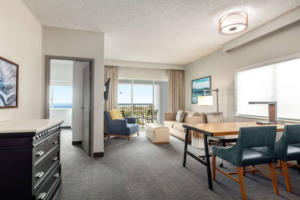 A hotel room with a large window, a sofa, a dining table, and a desk with drawers.