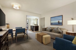 A hotel room with a flat-screen TV, a dining table with chairs, a sofa with patterned cushions, a coffee table, and a painting of a beach scene. - Embassy Suites by Hilton Monterey Bay Seaside (Seaside)