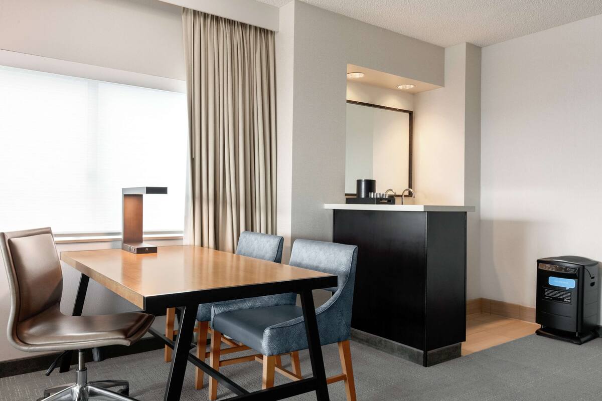 A hotel room with a wooden dining table, two chairs, a small black cabinet, a mirror, and a trash bin.