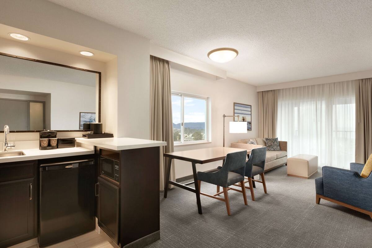A modern hotel room with a kitchenette, a dining area, a sofa, and a large window offering a view.