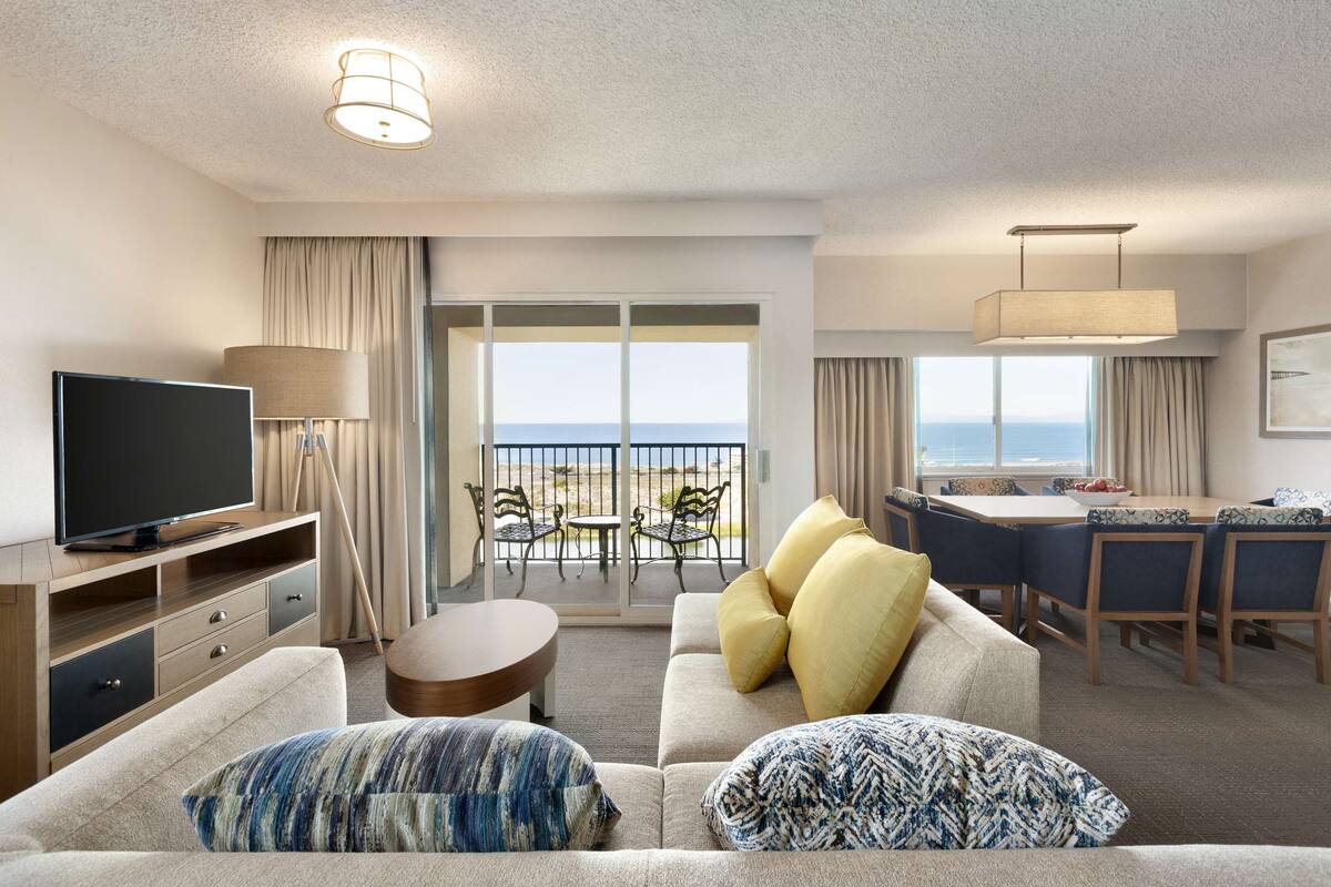 A hotel room with a sofa, TV, dining area, and a balcony with a view of the sea.