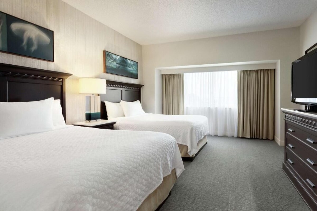A hotel room with two beds, a TV, a desk, and a window with curtains.