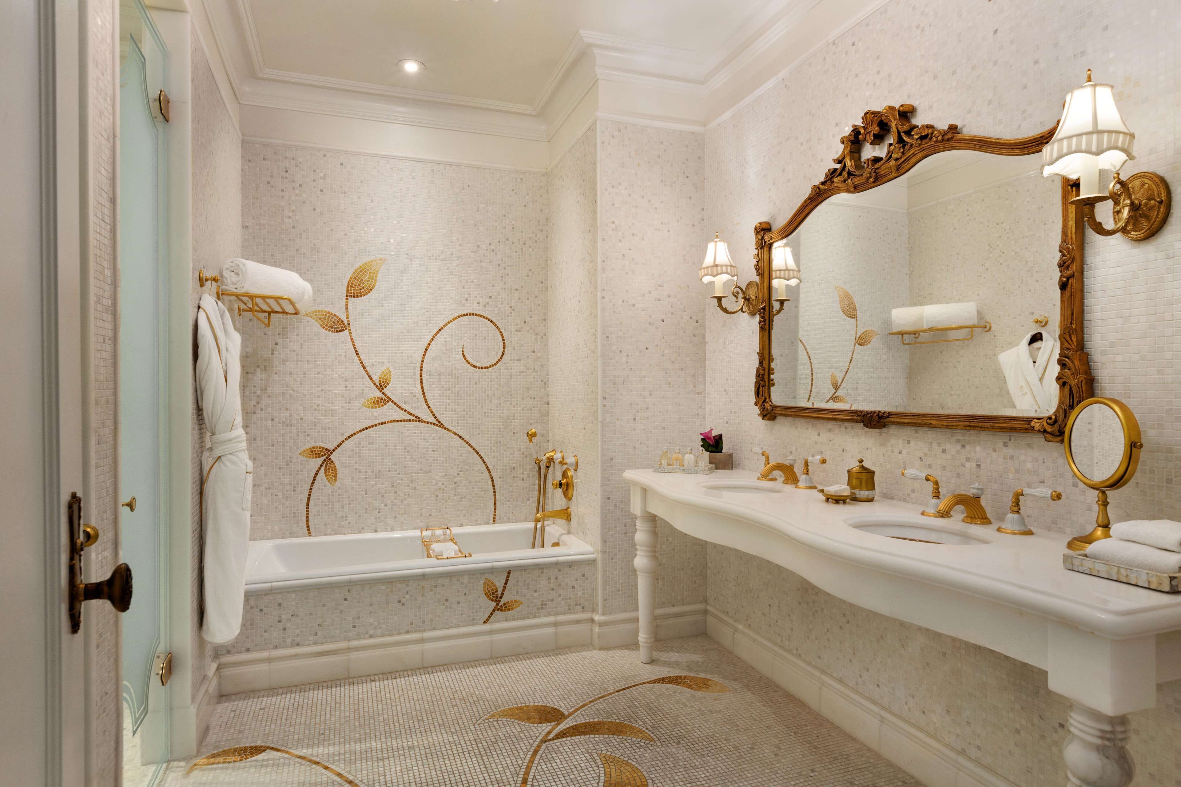 A luxurious bathroom with a white and gold color scheme, featuring a freestanding bathtub, a large mirror, and a vanity with a sink and towel rack.